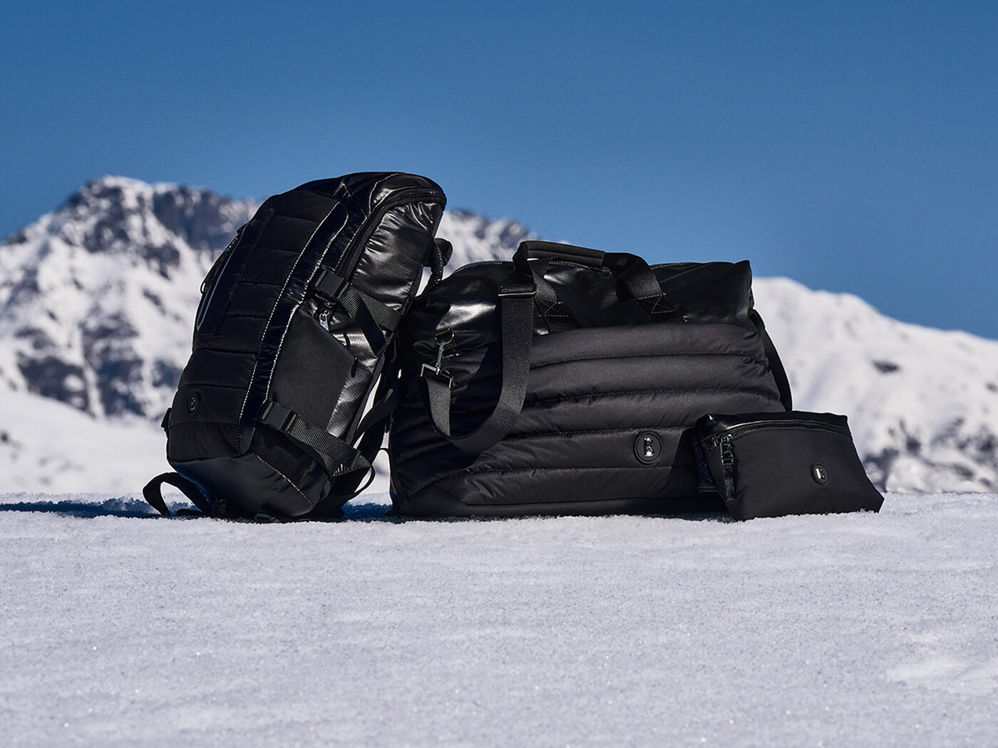 The image shows three different black bags in the snow arranged in front of a backdrop of mountains. 
