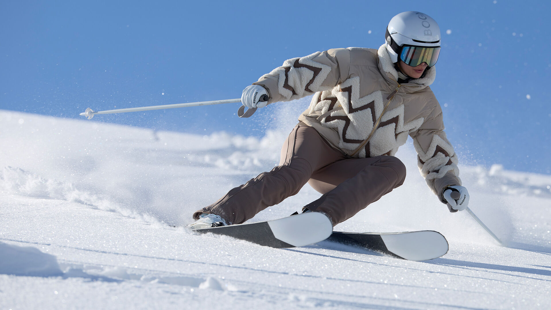 The image shows a model skiing down a freshly groomed slope. She is wearing a ski jacket with a zigzag pattern and brown ski pants.