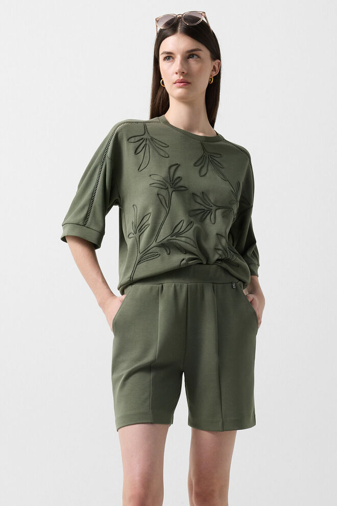 Das Damen Model tr&auml;gt einen Komplettlook bestehend aus einem Kurzarm-Sweat und Shorts in Oliv-Gr&uuml;n mit Broderie Anglaise und floralen Stickereien.