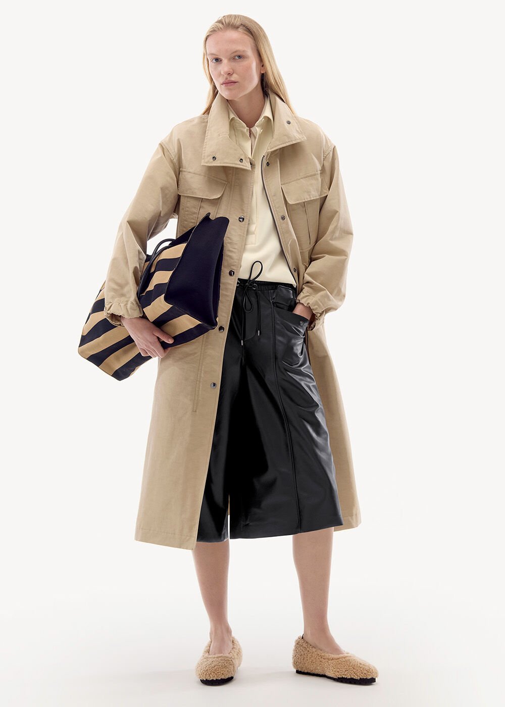 This model is wearing a beige trench coat with patch pockets, a black leather skirt and shearling ballerina flats.