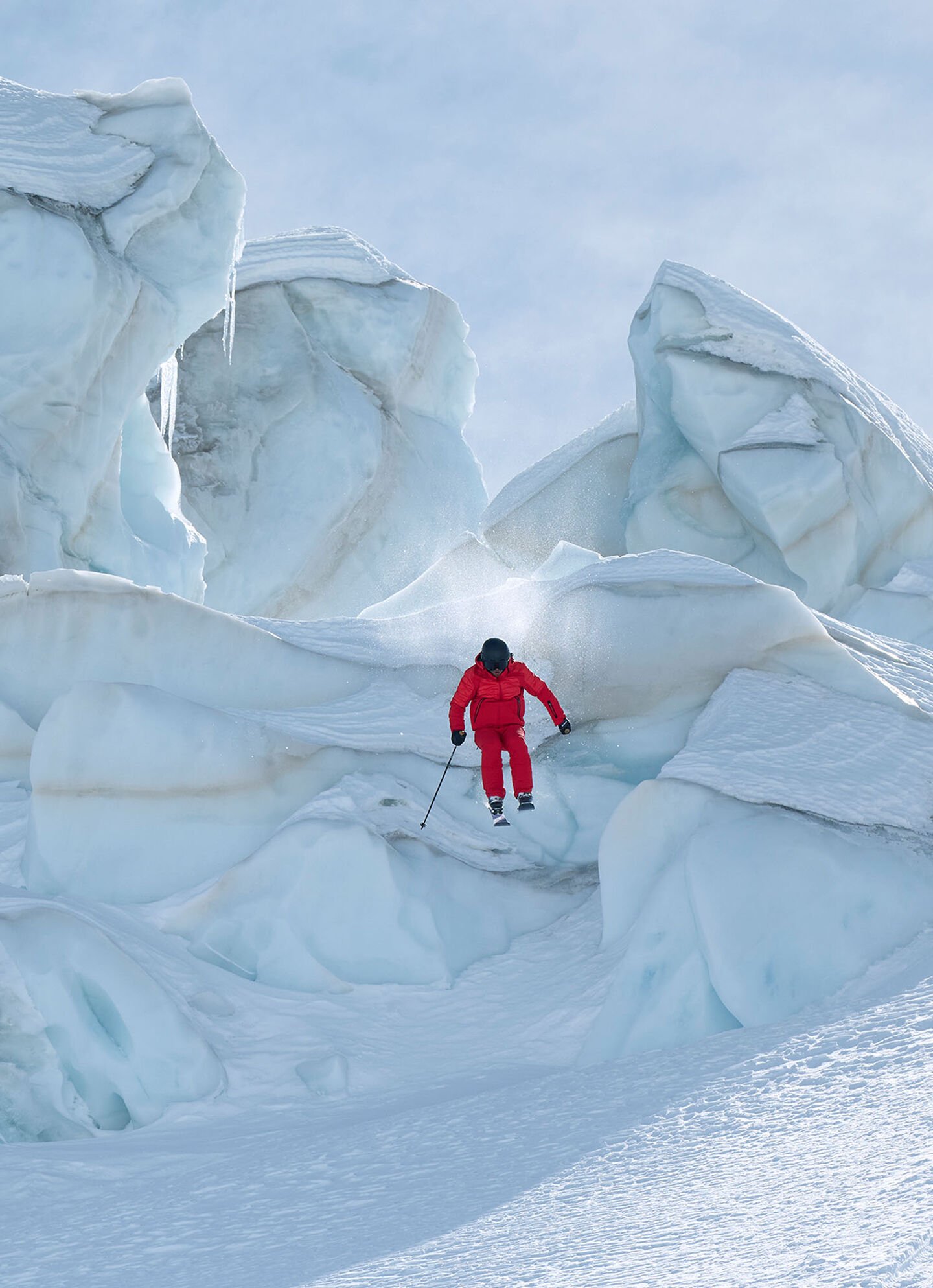 Ein Skifahrer springt von einem Skiabhang herunter - bekleidet in roter Skijacke und Skihose.