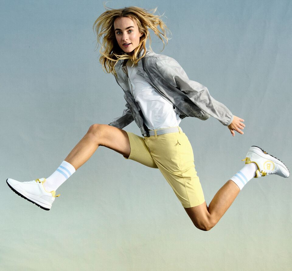 The blonde model strikes a dynamic pose wearing a polo shirt, a printed blouson jacket, yellow shorts and golf trainers.