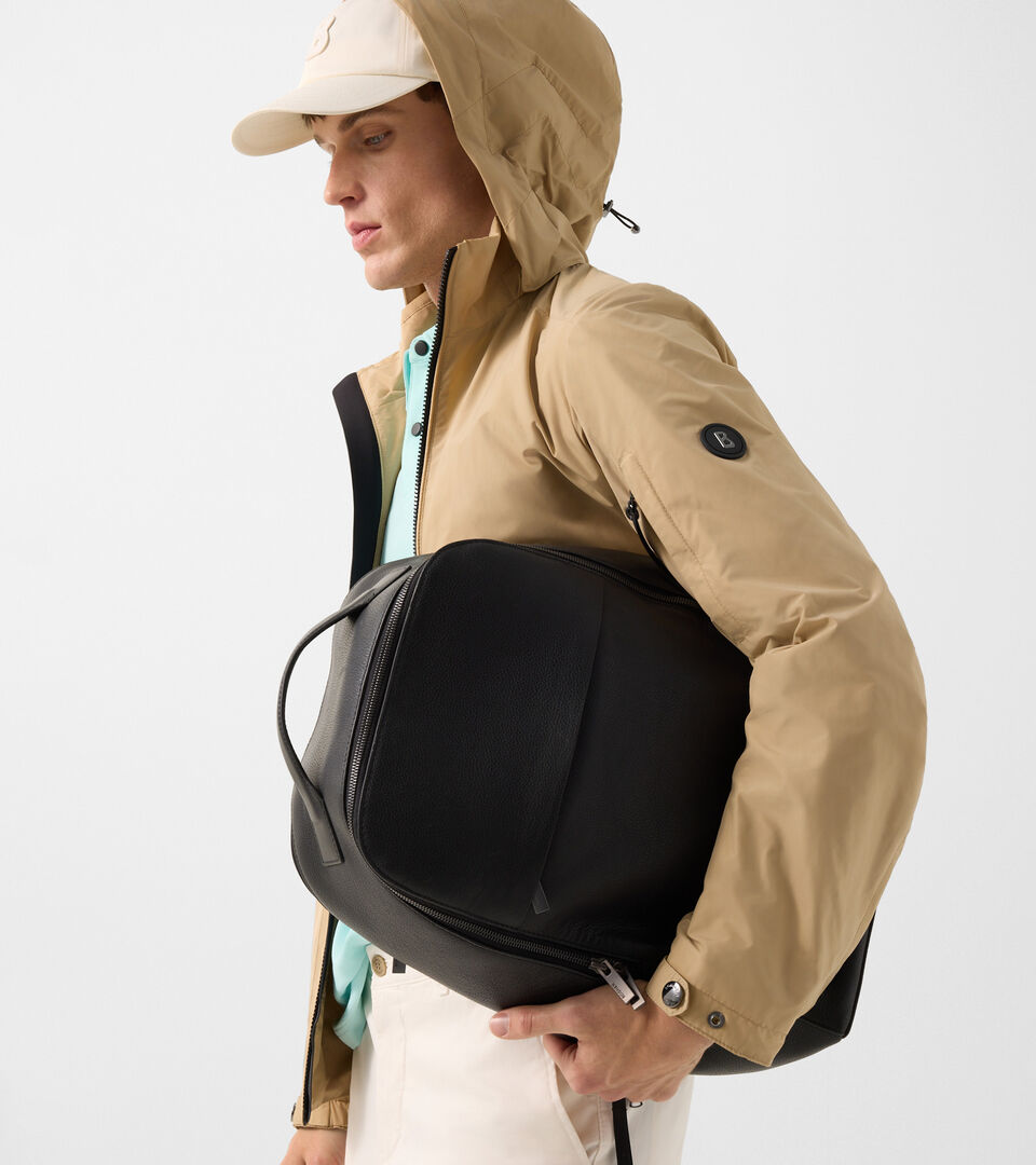 Seitliche Aufnahme des Herren Models mit heller Jacke und Polo-Shirt. Er h&auml;lt einen schwarzen Rucksack im Arm.