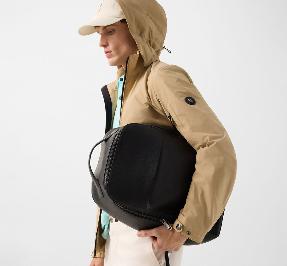 Seitliche Aufnahme des Herren Models mit heller Jacke und Polo-Shirt. Er h&auml;lt einen schwarzen Rucksack im Arm.