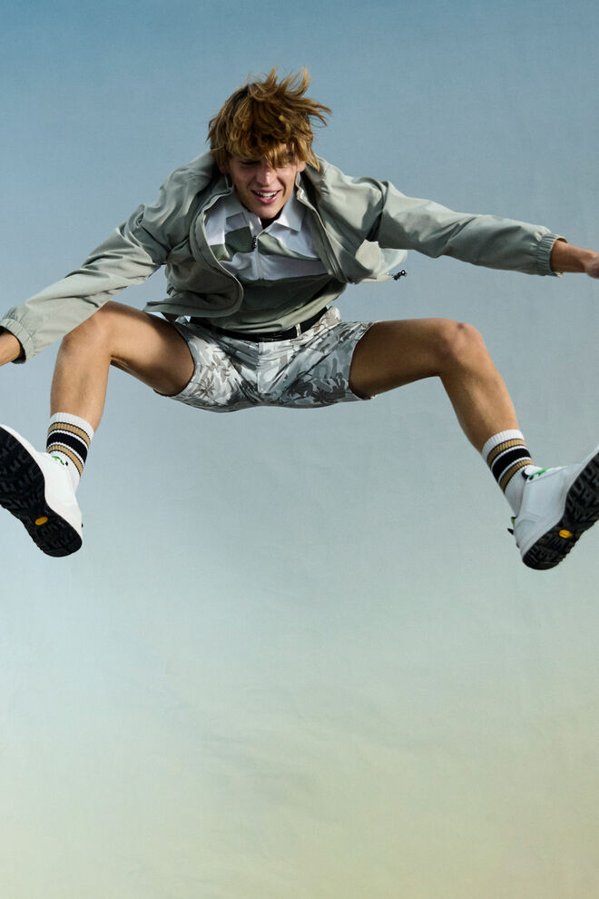 The male model is seen mid-jump. He wears a lightweight blouson jacket in eucalyptus green and palm-printed shorts.