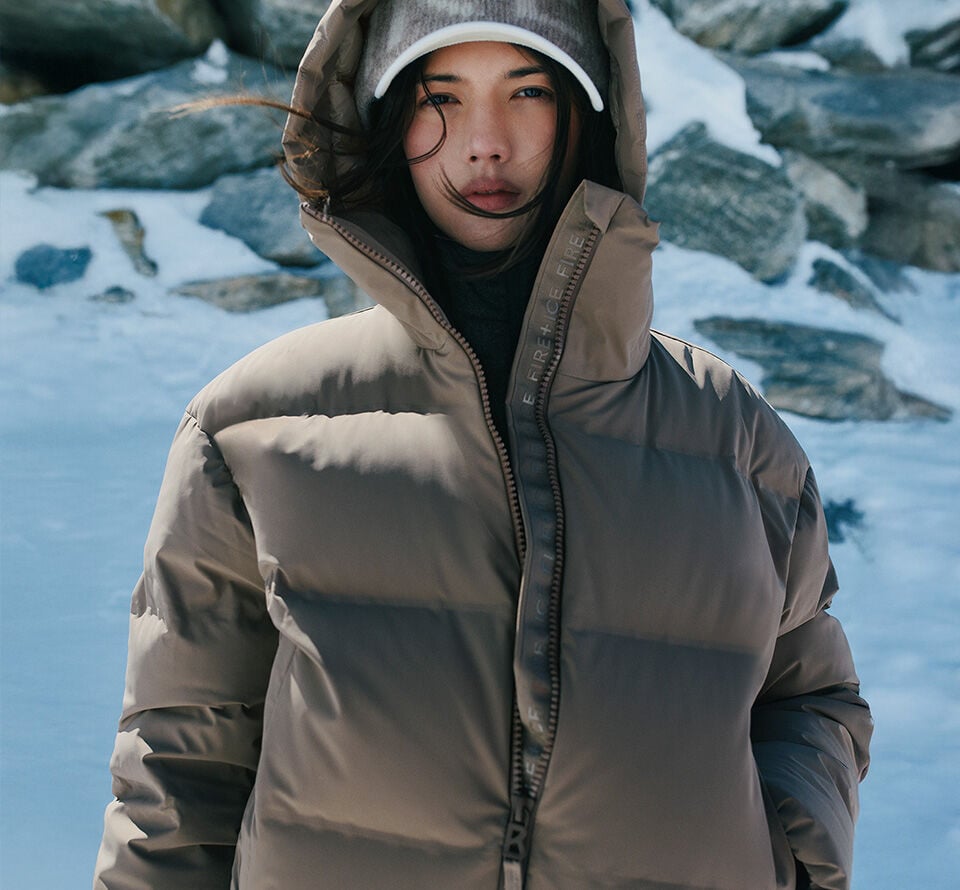 Das Damen Model trägt eine kurze karamellfarbene Daunenjacke mit Kapuze und Logo-Borte am Reißverschluss.