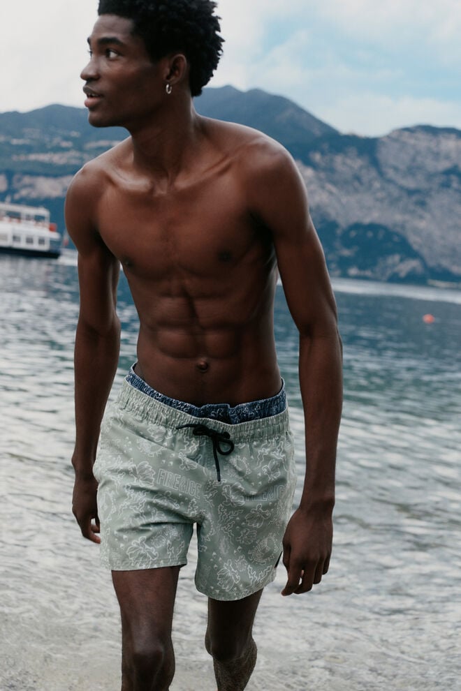 The male model is wearing blue and green swimming trunks in a striking pattern with a FIRE+ICE logo print. There is a mountain lake in the background.