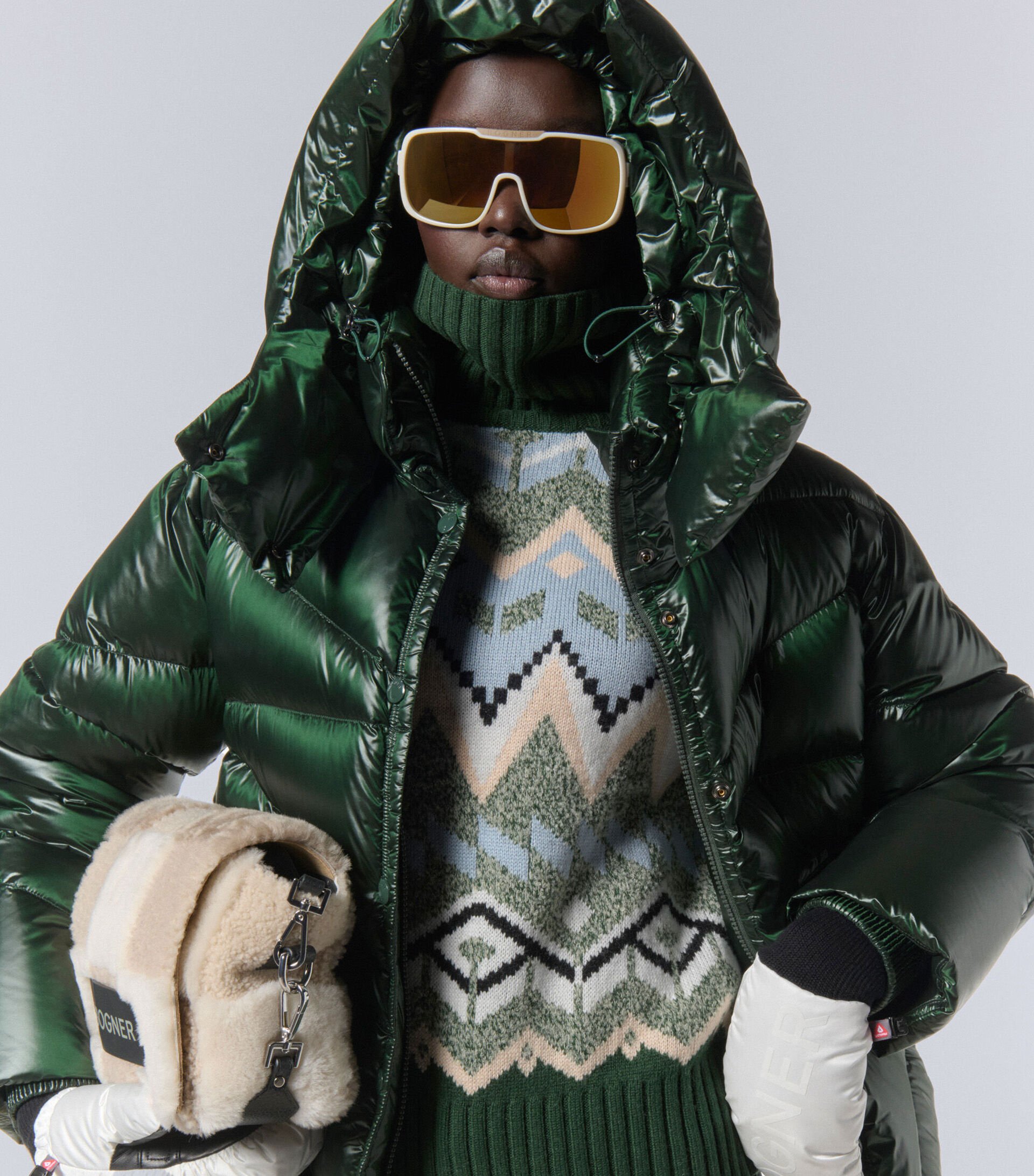 Close-up vom Damen Model. Sie trägt eine dunkelgrüne, glänzende Daunenjacke, einen Norwegerpullover und eine Sonnenbrille.