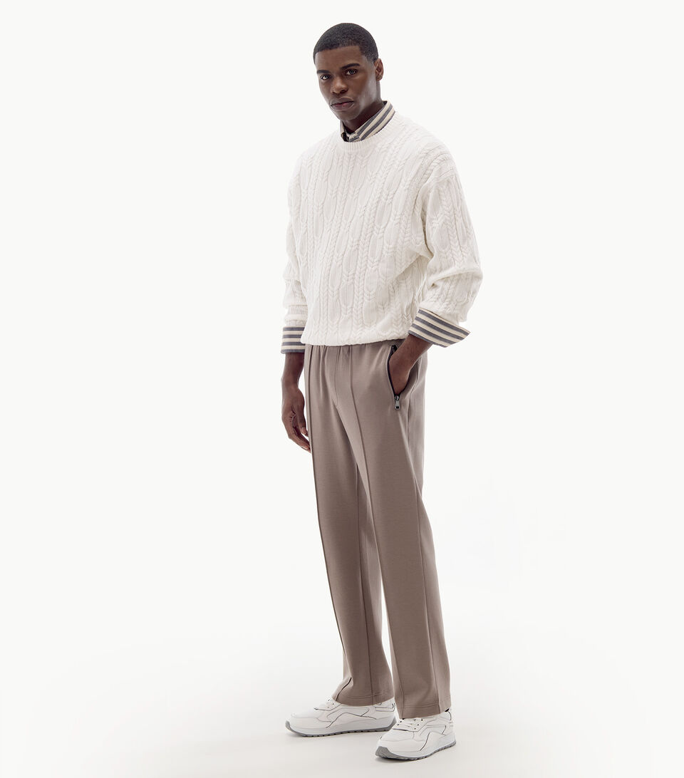 Ganzkörperaufnahme des Herren Models mit gestreiftem Hemd, darüber ein Zopfstrick-Pullover und schlammfarbene Sweatpants.