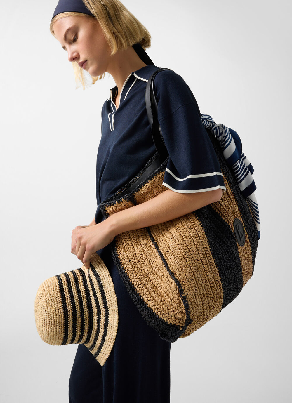 The blonde female model in navy blue clothes carries a big tote bag made from natural fibres and a matching hat.