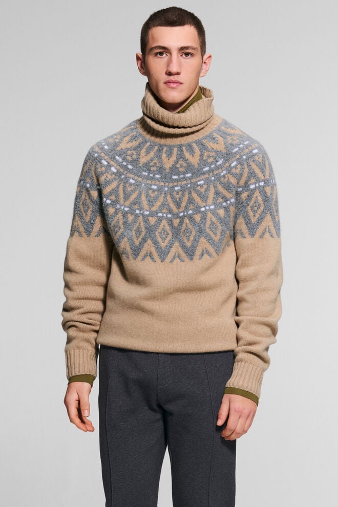 The male model is wearing the Roxter Fair Isle cashmere sweater in Camel with a grey pattern, paired with wool trousers.