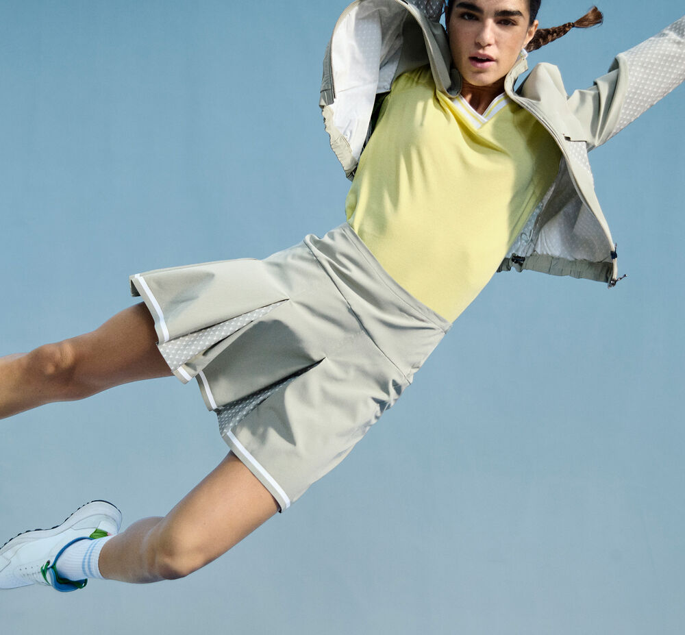 The dark-haired model is photographed mid-jump. She wears a bright yellow polo shirt, a light green blouson jacket and a golf skirt.