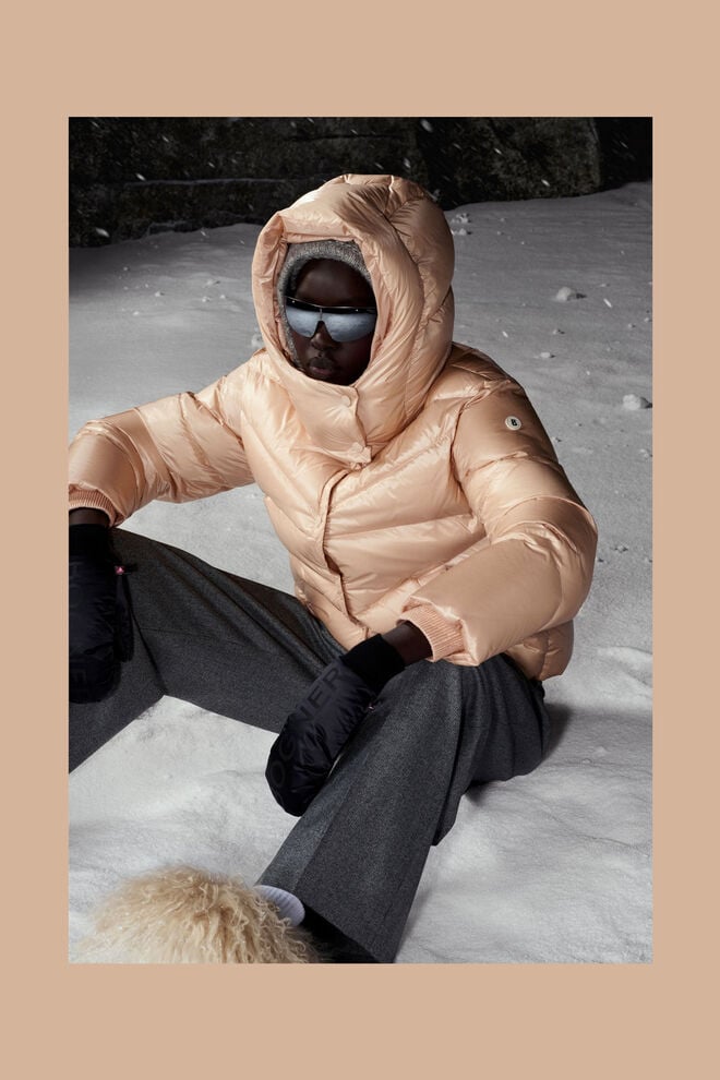 Das Model sitzt auf schneebedecktem Boden und trägt Daunenjacke Xally in hellem Apricot.
