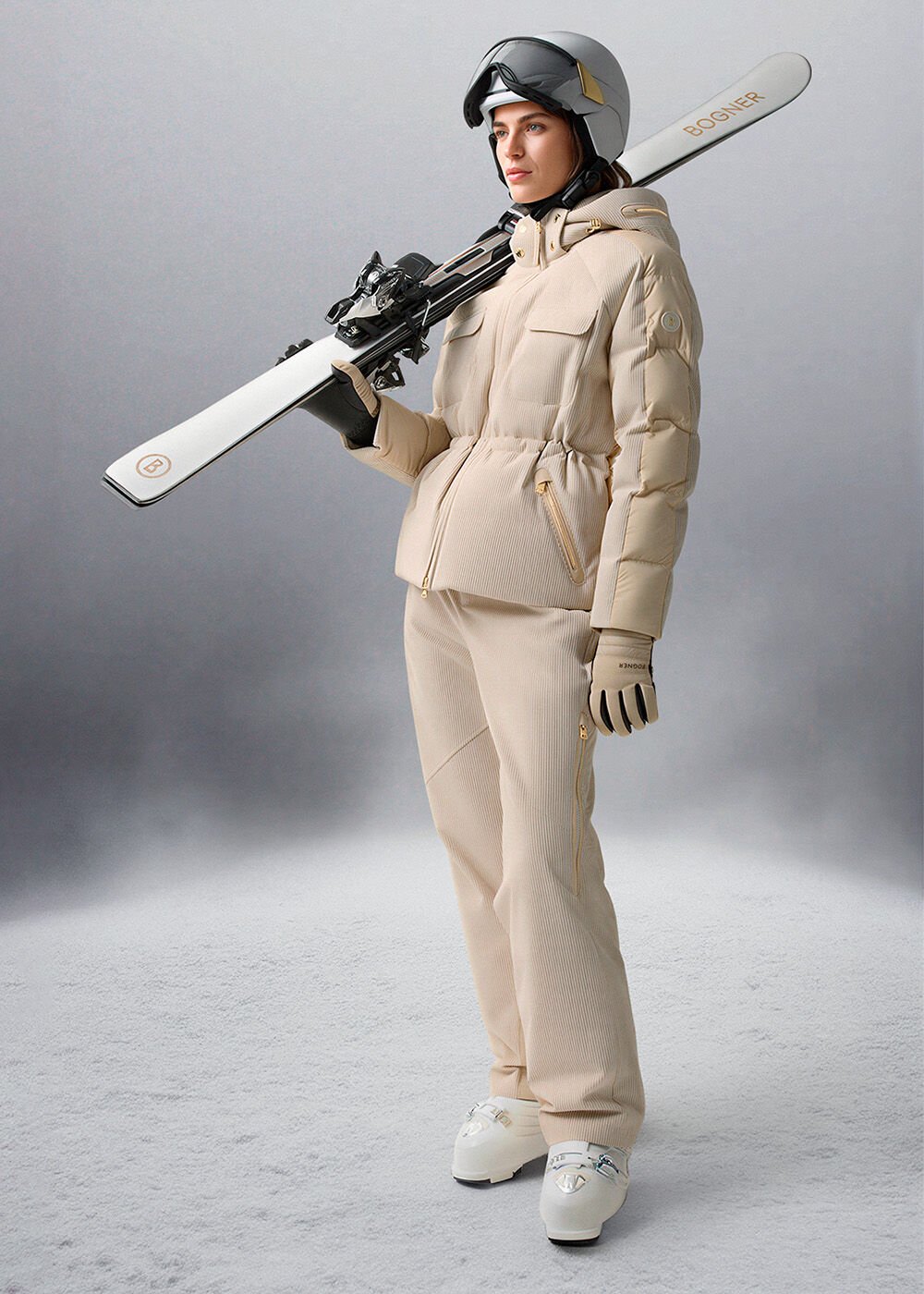 Das Damen-Model trägt ein beigefarbenes Ski-Outfit, einen weißen Skihelm und hält Ski in den Händen.