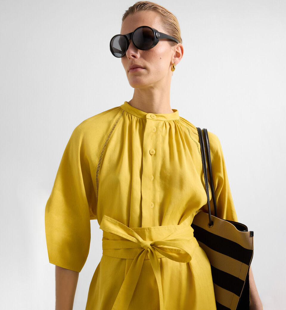 Das blonde Damen Model tr&auml;gt ein Kleid in Sonnengelb, das mit fein gearbeiteten Broderie Anglaise Details bestickt ist.