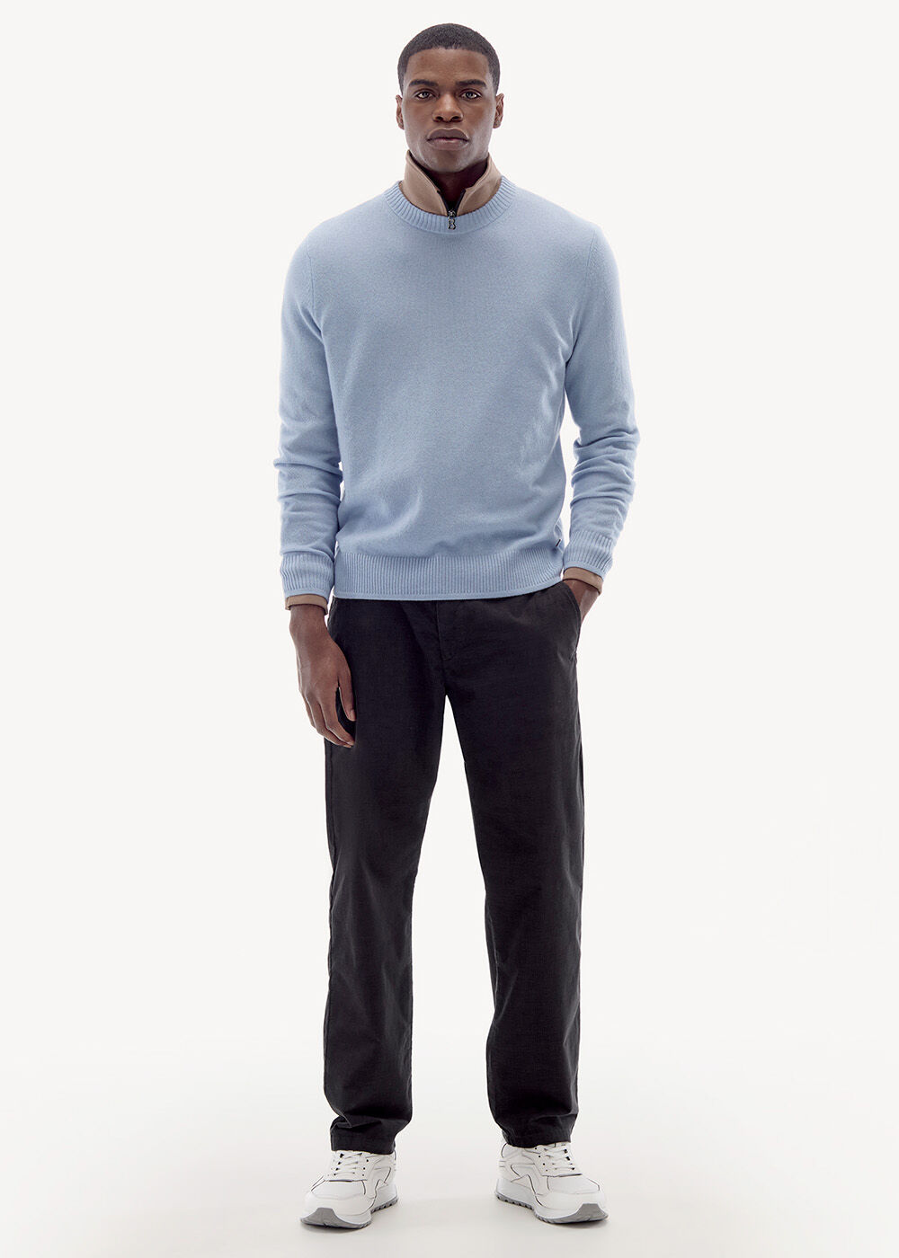 Dieser Look besteht aus einem hellblauen Pullover, darunter ein hellbrauner Troyer. Dazu dunkle Jeans und weiße Sneaker.