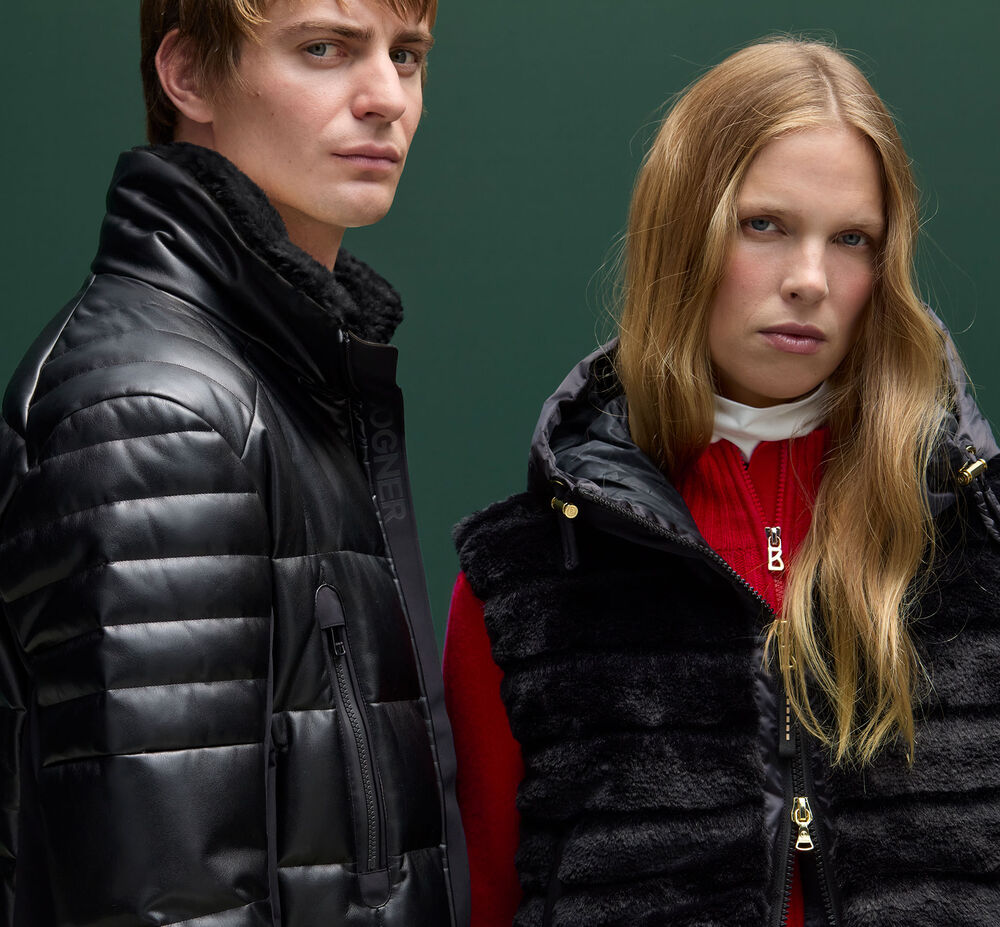 Close-up of a man and a woman in a black jacket and gilet standing in front of a fir-green background.