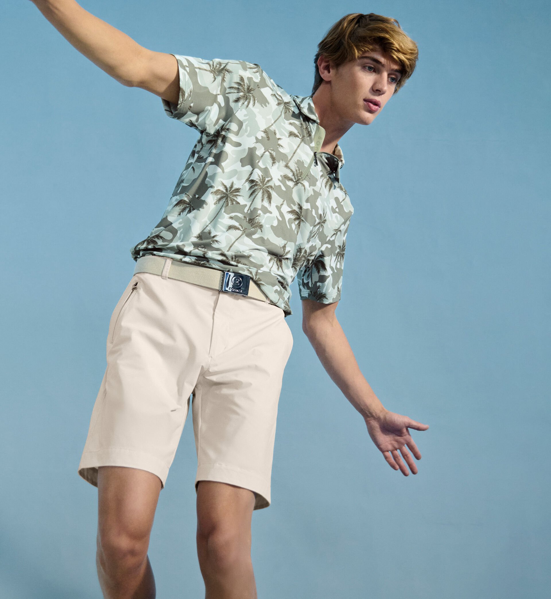 Das blonde Herren Model in dynamischer Pose tr&auml;gt ein Polo-Shirt mit Palmen- und Camouflage-Print, dazu eine Shorts und einen G&uuml;rtel.