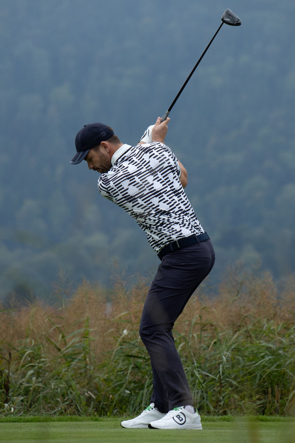 Seitliche Aufnahme des Herren Models beim Abschlag. Er tr&auml;gt ein Polo-Shirt mit grafischem B-Print und eine dunkle Hose.