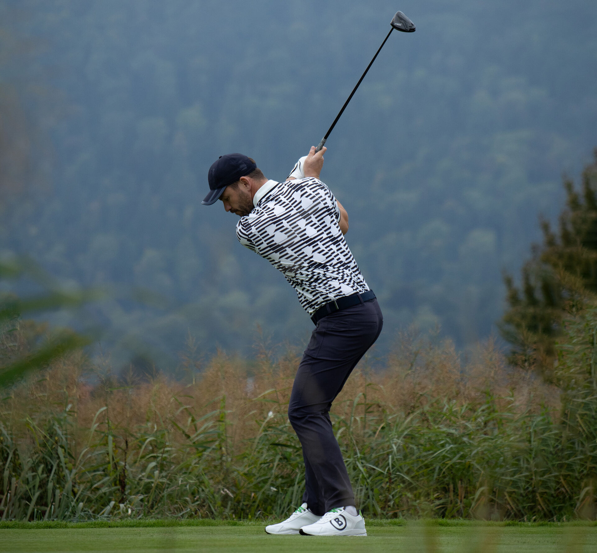 Seitliche Aufnahme des Herren Models beim Abschlag. Er tr&auml;gt ein Polo-Shirt mit grafischem B-Print und eine dunkle Hose.