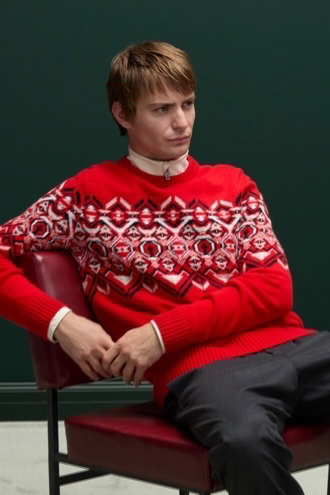 Le mannequin masculin assis porte le pull norvégien Luci dans un rouge éclatant. On distingue en dessous un col roulé avec fermeture éclair en B.