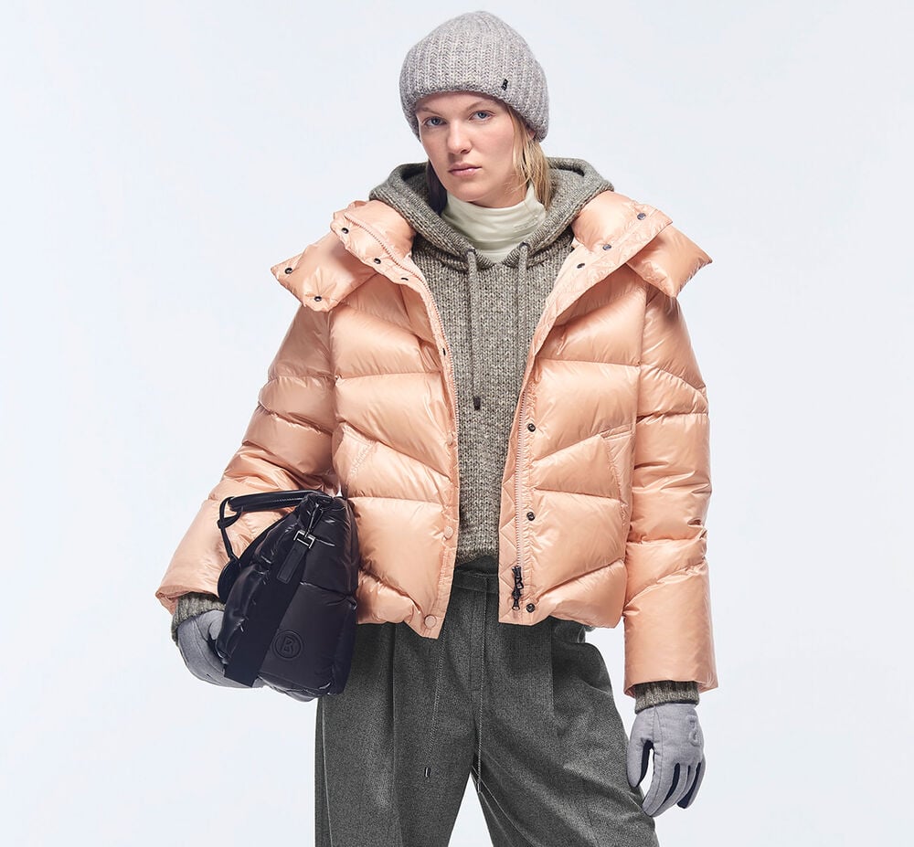 The model is wearing a cropped down jacket in apricot and a woolly hat, knitwear and pants in grey with a black bag.