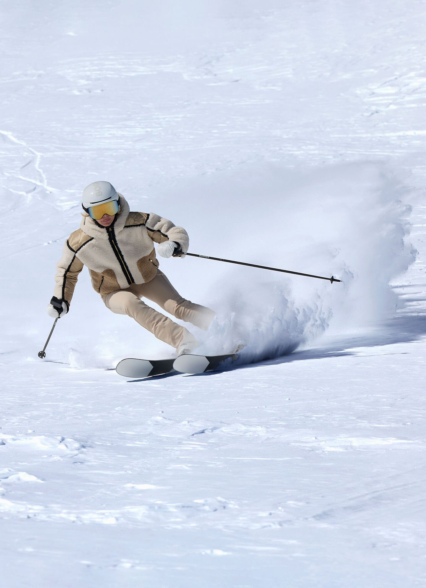 Das Model fährt schwungvoll eine Skipister herunter und trägt dabei eine beige Skijacke, Skihose und einen Skihelm.