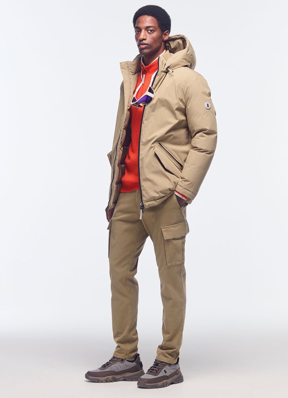 Het model draagt een dikke beige donsjas, een rode troyer en een camelkleurige cargobroek. 