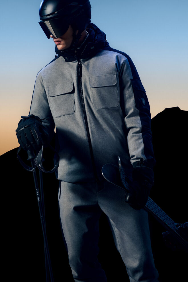 Seitliche Ganzk&ouml;rperaufnahme. Im Hintergrund eine Bergkulisse. Das Herren Model tr&auml;gt Ski-Daunenjacke Boyen aus Tech-Wolle.
