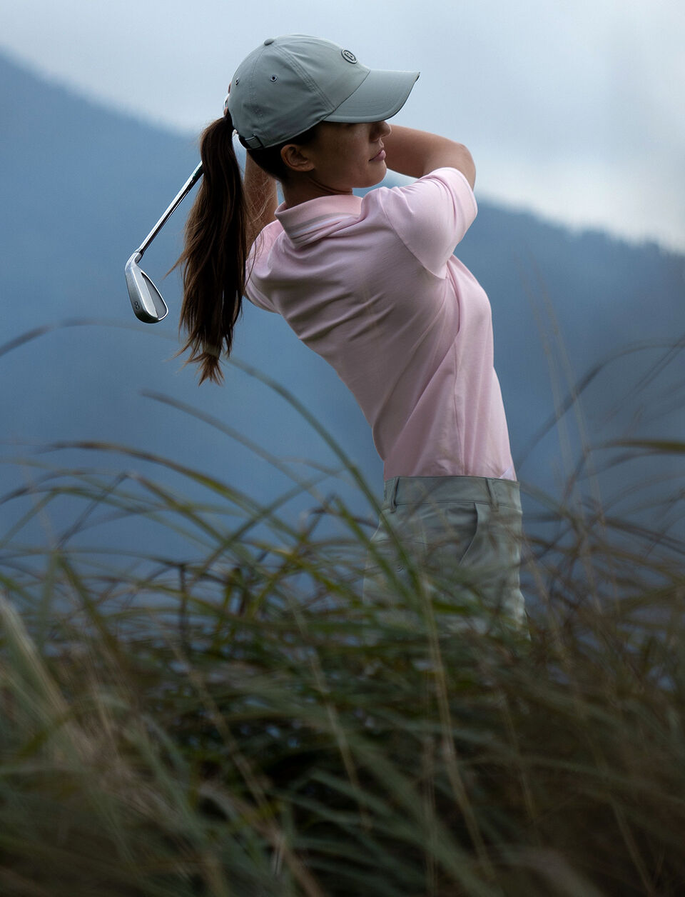 The female golf player is seen from the side. She is teeing off and wears a light pink polo shirt, a base cap and green pants.