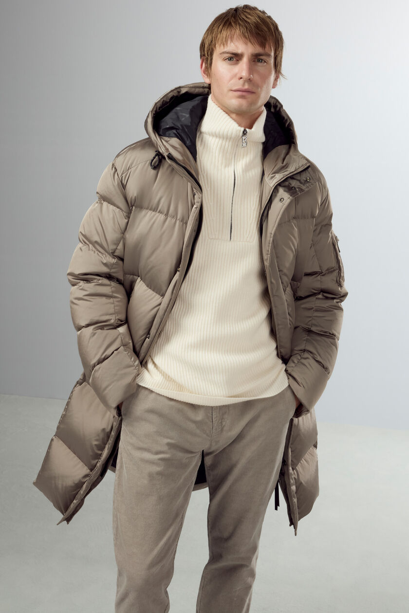 Das Herren Model trägt einen cremefarbenen Kaschmir-Troyer in Rippstrick, dazu einen Daunenmantel und helle Jeans.