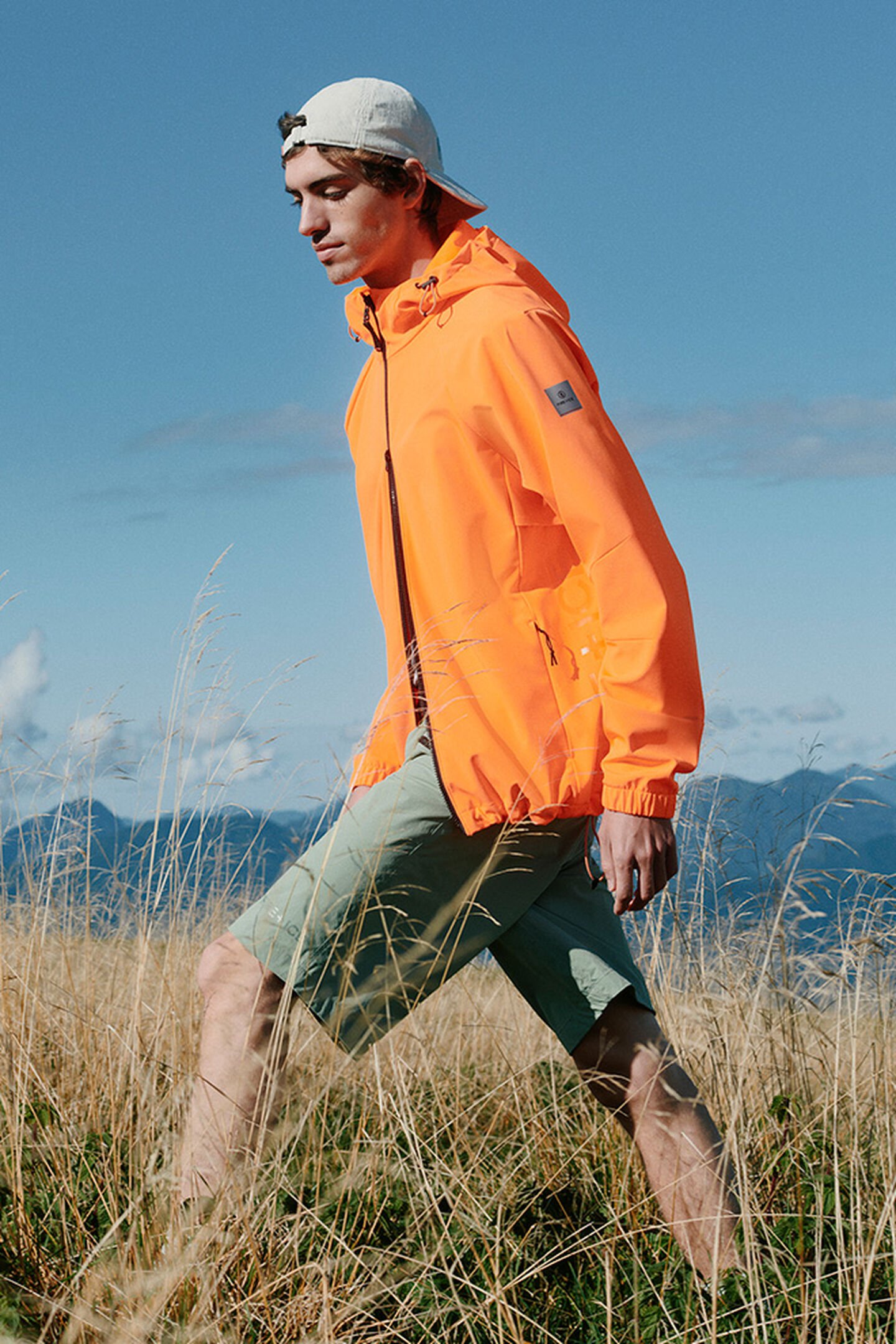 The male model hiking through the meadows is wearing a performance jacket in bright orange with green shorts.