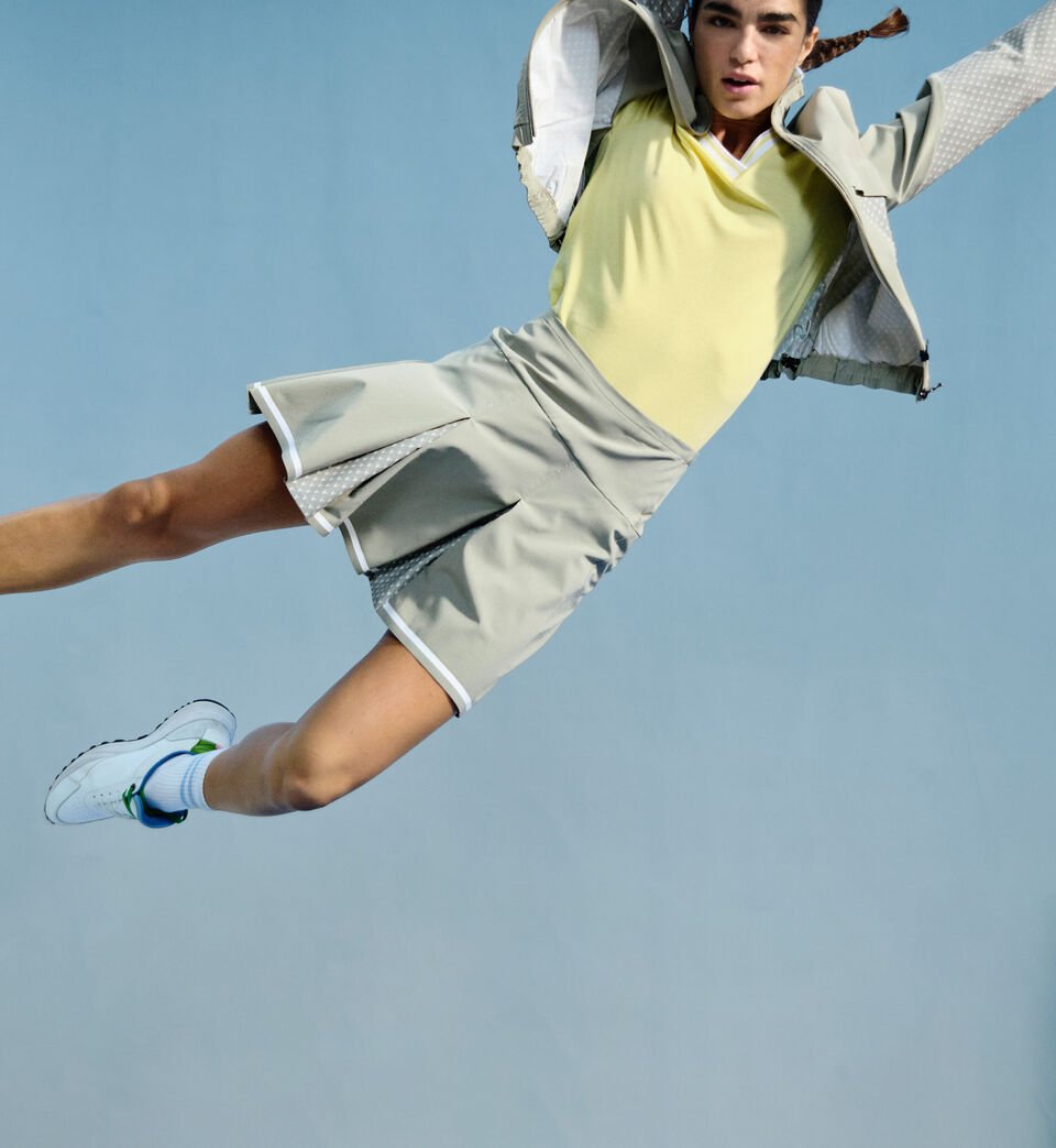 Das blonde Damen Model tr&auml;gt ein Polo-Shirt mit Logo-Zip, dazu eine gelbe Hose und einen leichten Blouson mit Palmen-Print.