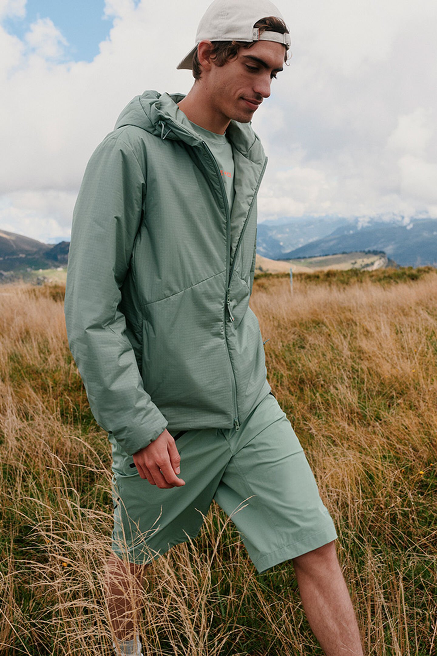 The male model is wearing a set in eucalyptus &ndash; jacket and shorts &ndash; and is hiking through the meadows of a mountain landscape.
