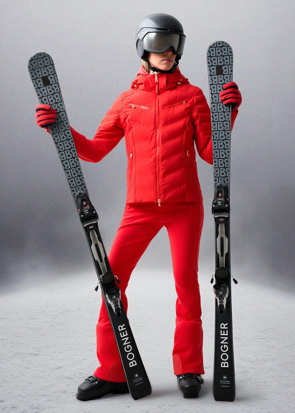 Das Herren-Model trägt ein rotes Ski-Outfit, einen schwarzen Skihelm und hält Ski mit B-Print in den Händen.