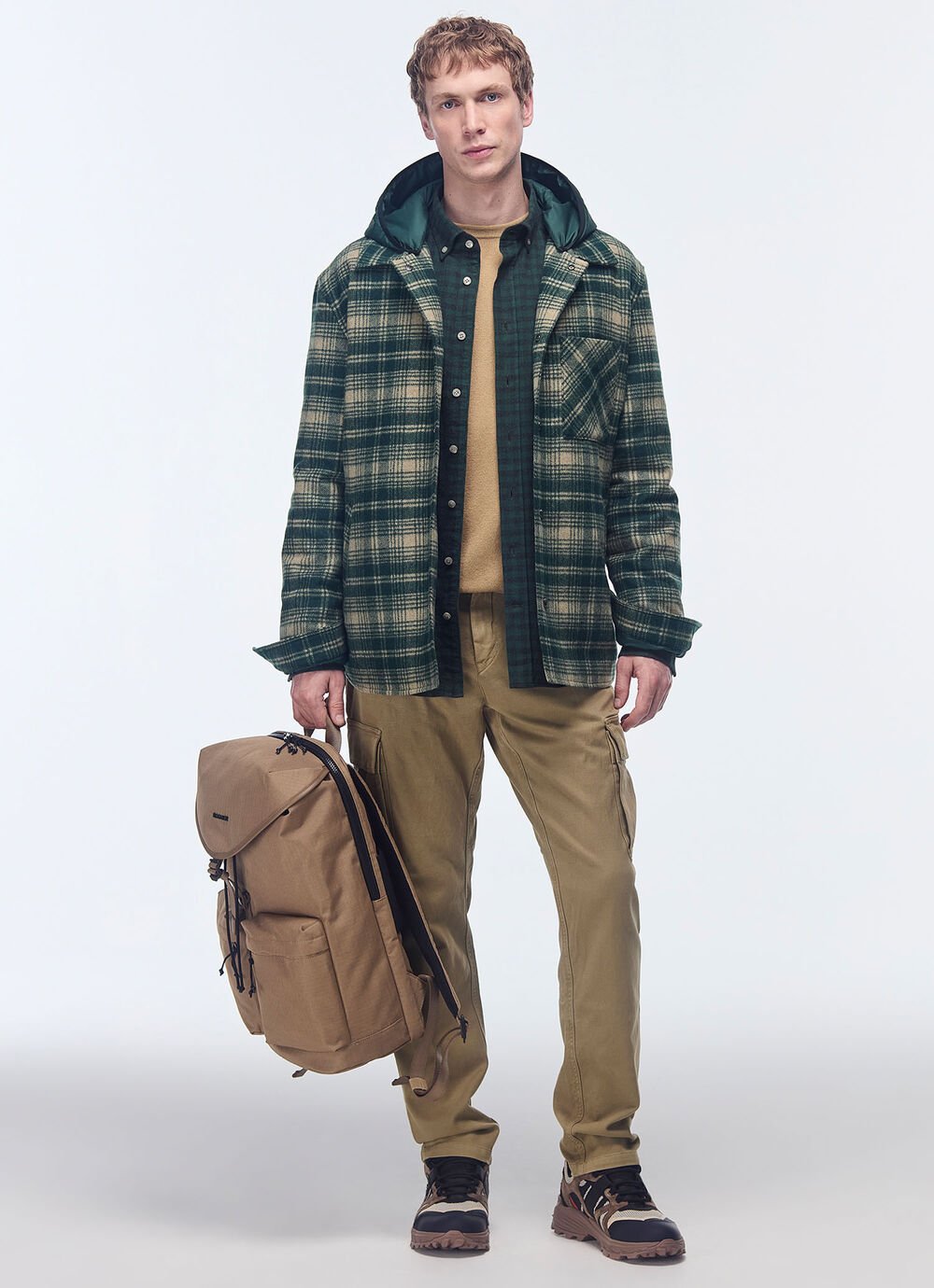 Het model draagt een groen/beige geruit overhemdjasje met een satijnen capuchon, samen met een trui en een camelkleurige cargobroek. 
