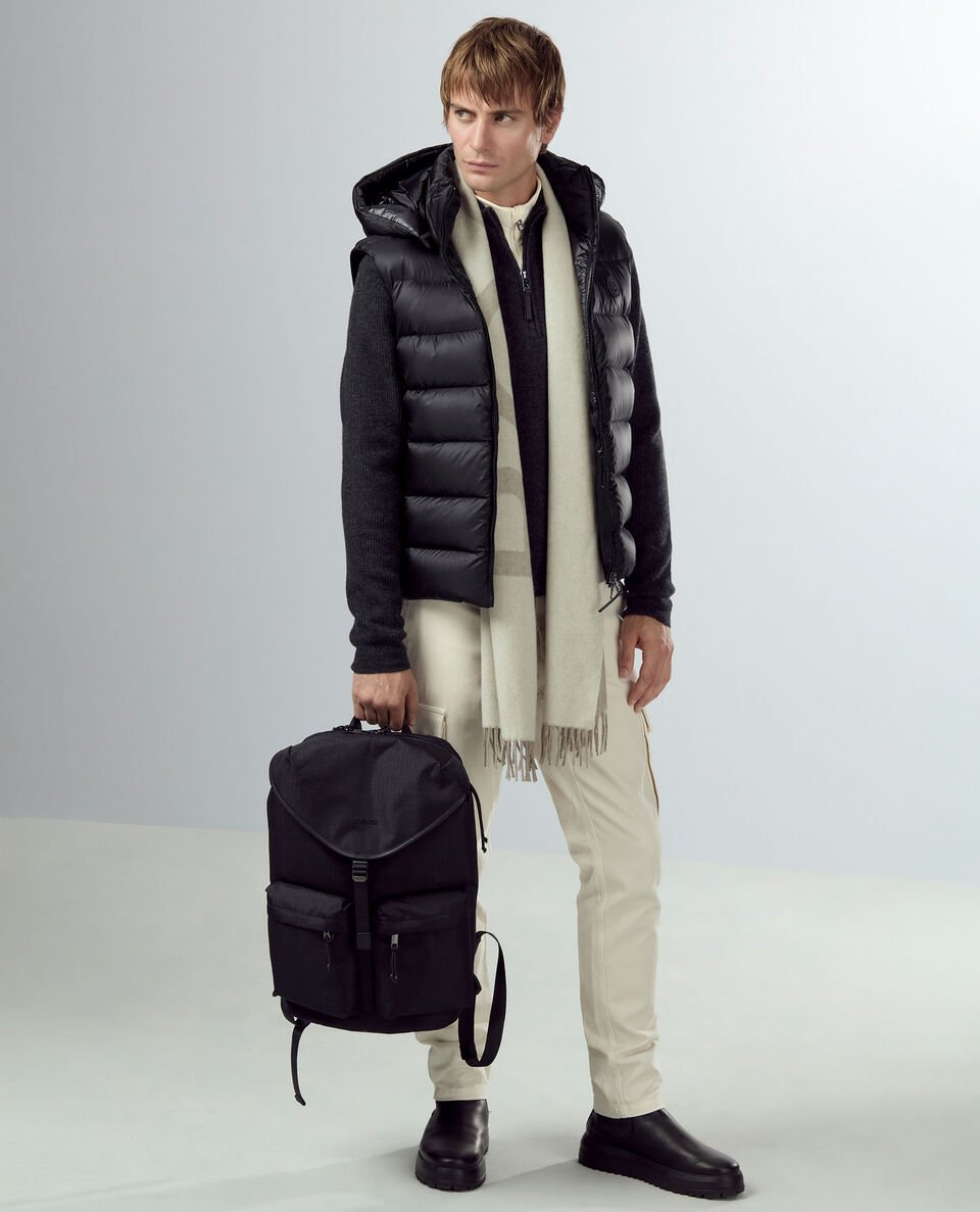 Das Herren Model trägt eine schwarze Daunenweste mit gemustertem Schal, eine helle Hose und einen Rucksack in der Hand. Das Herren Model trägt eine schwarze Daunenweste mit gemustertem Schal, eine helle Hose und einen Rucksack in der Hand.