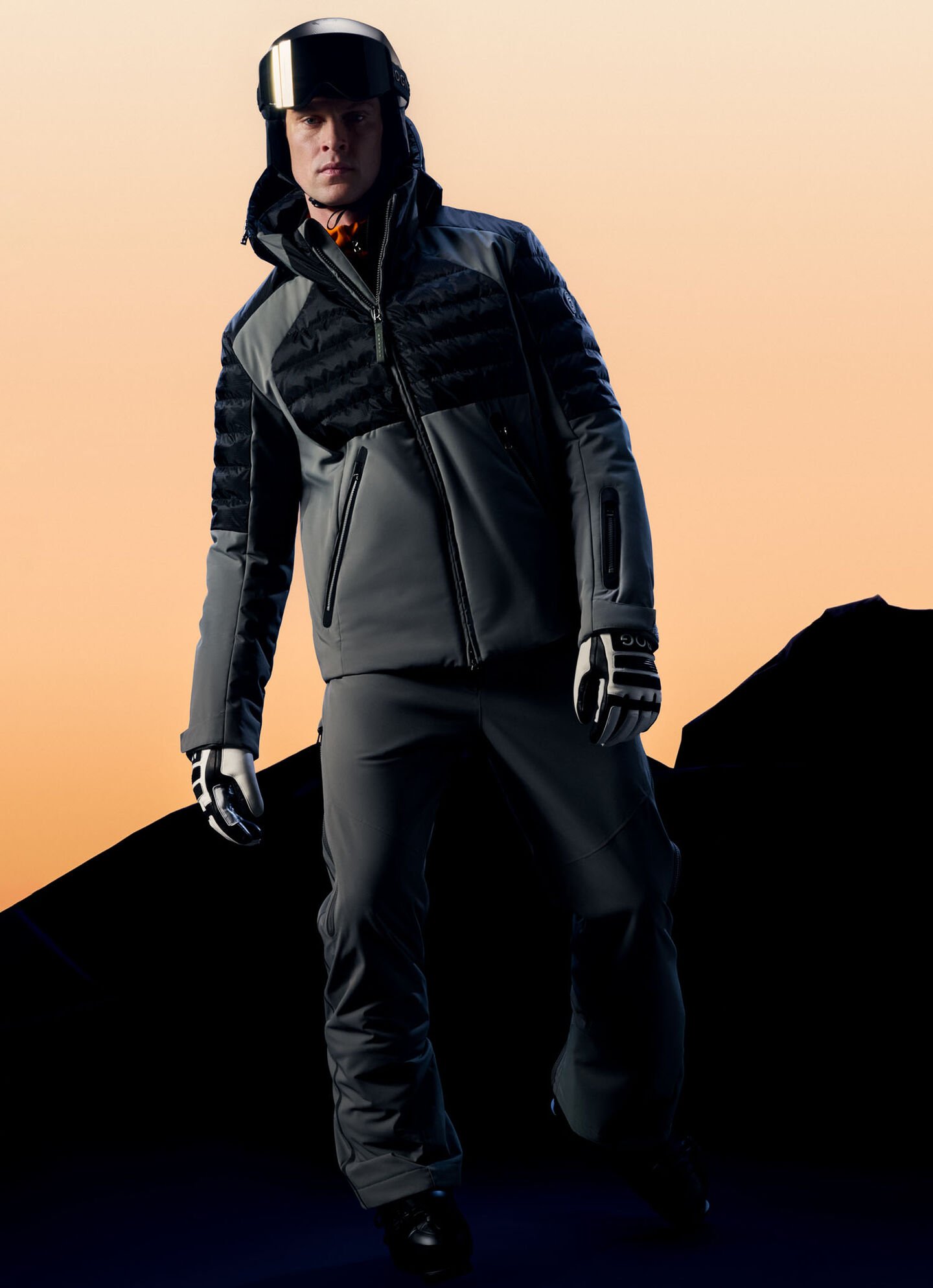 Das Model trägt eine graue Skijacke mit gepaddeten Einsätzen, eine graue Skihose, einen Skihelm und Skihandschuhe.