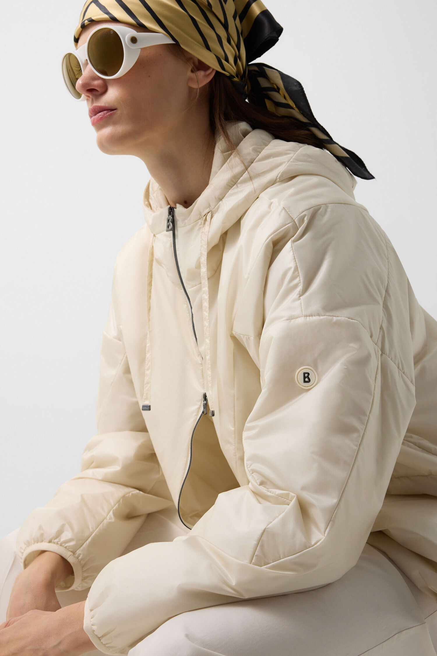 Side view of the female model wearing a cream-coloured Beryll lightweight jacket. She is also wearing a scarf and sunglasses.
