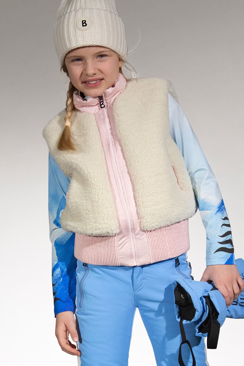 A zip-up gilet complete with teddy fur and a white woolly hat complement this girl&rsquo;s look, which comprises a first layer and ski trousers in blue.