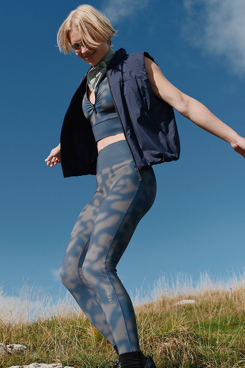 Ein Damen Model in dynamischer Pose in einer grasbedeckten Landschaft. Sie tr&auml;gt ein Set aus Zip-Top und Tights mit Print.