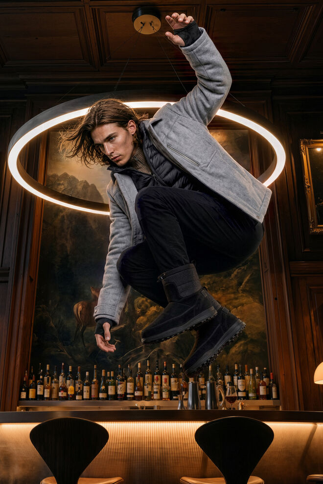 Men's model in a dynamic pose in front of a hotel bar wearing the grey Flave flannel jacket with a black quilted inlay, paired with black trousers and winter boots.