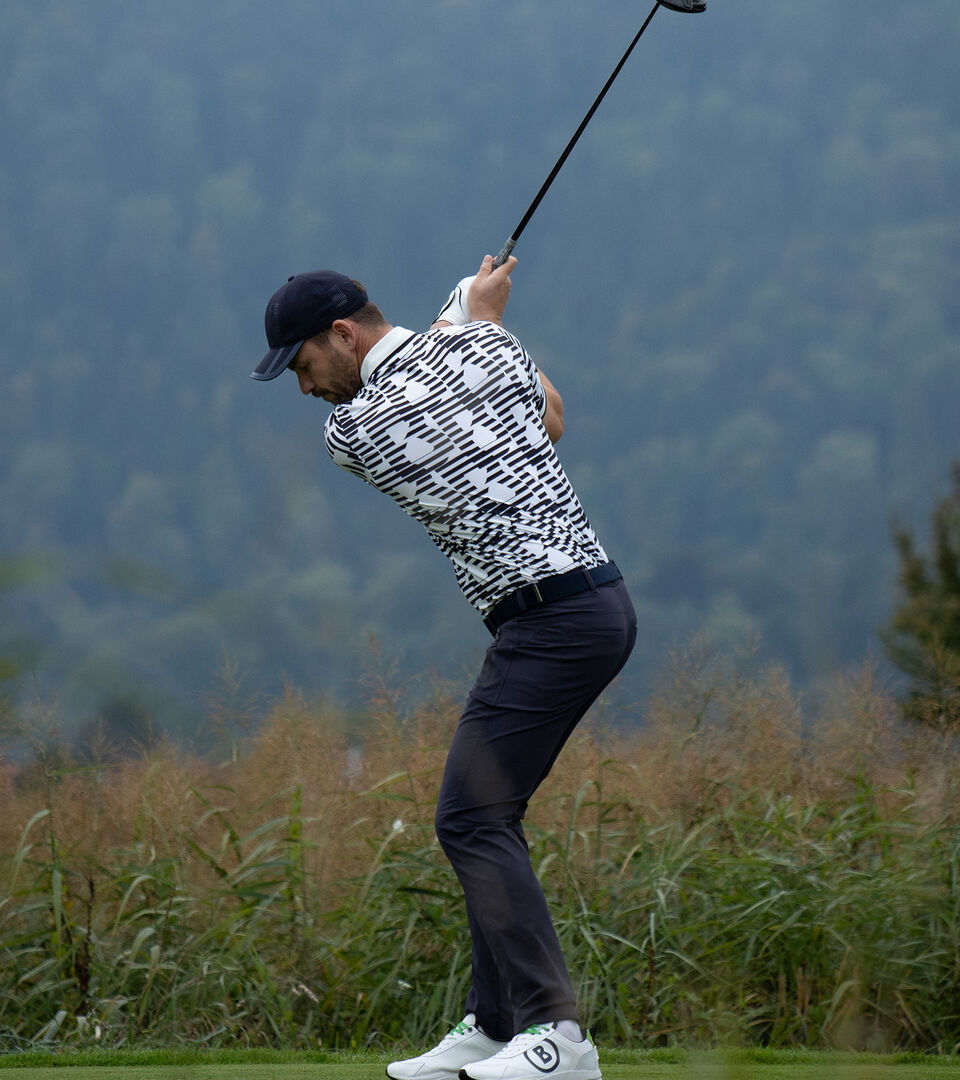 Side view of the male golf player while teeing off. He wears dark blue long pants and a polo shirt with distinctive &lsquo;B&rsquo; print.