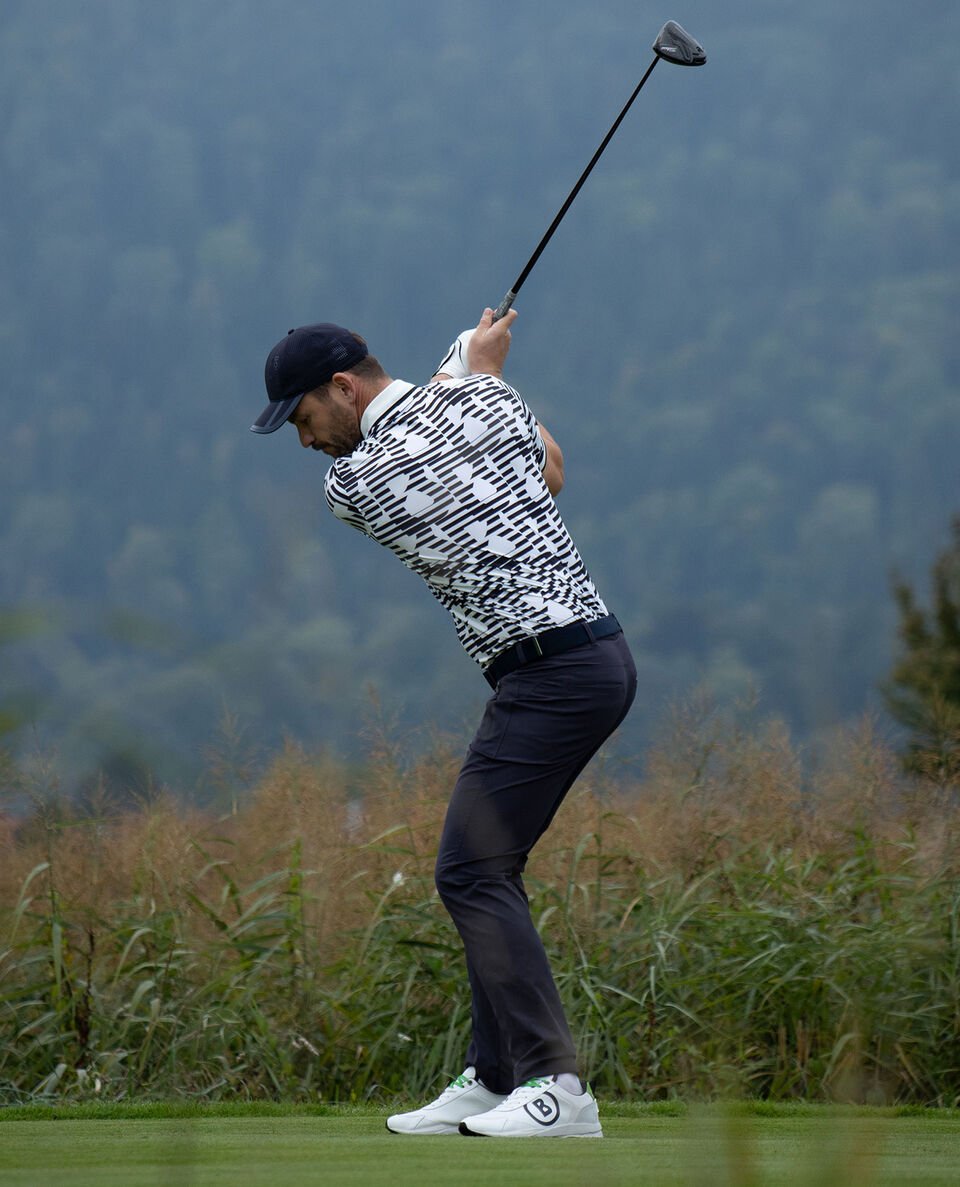 Side view of the male golf player while teeing off. He wears dark blue long pants and a polo shirt with distinctive &lsquo;B&rsquo; print.