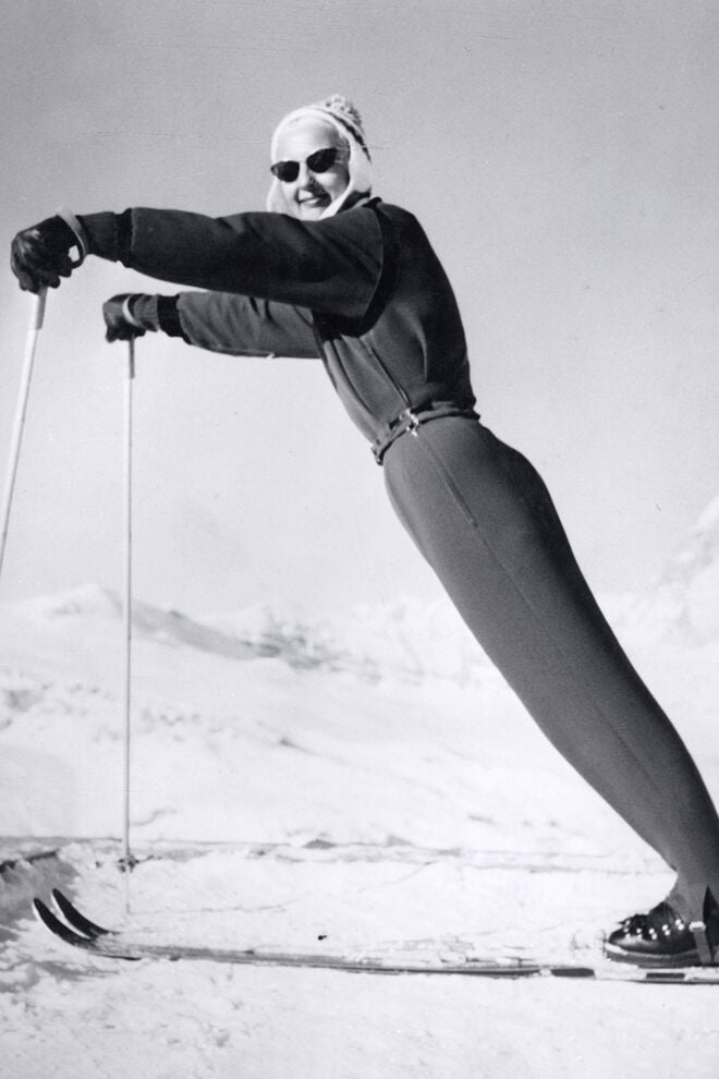 Maria Bogner striking a sporty pose at the summit. She is wearing a ski outfit comprising a short jacket, stirrup trousers, a knitted hat and skis.