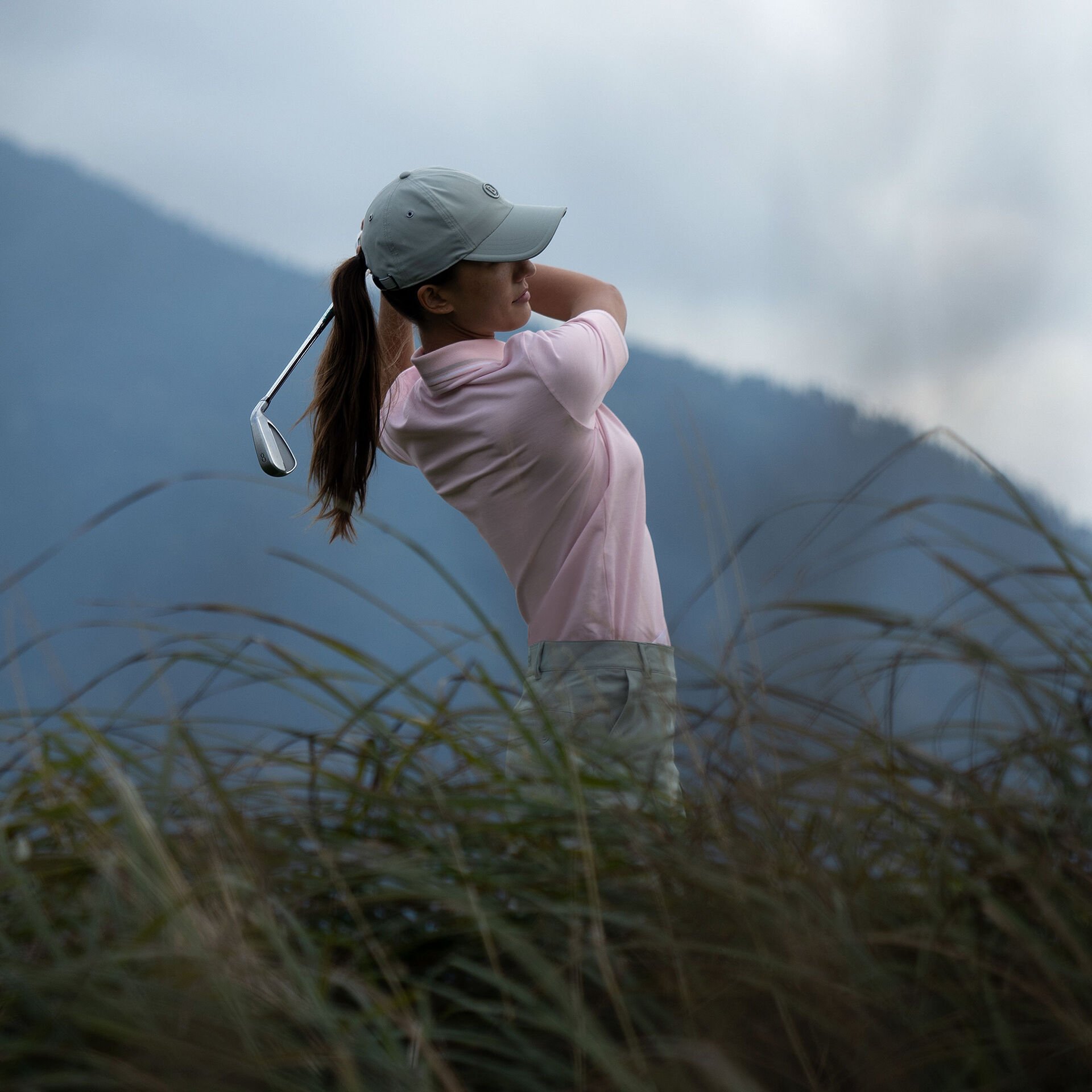 A side view half-body shot of a female golfer teeing off wearing a pink polo shirt. Mountain scenery behind.