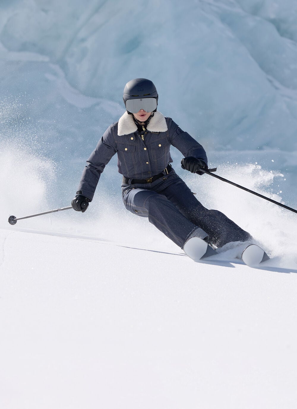 Das Damen Model in Action auf der Piste tr&auml;gt Ski-Overall Jenna aus Denim mit kontrastierendem Fellkragen, dazu Skist&ouml;cke und Skier.