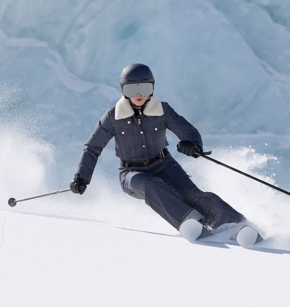 Das Damen Model in Action auf der Piste tr&auml;gt Ski-Overall Jenna aus Denim mit kontrastierendem Fellkragen, dazu Skist&ouml;cke und Skier.