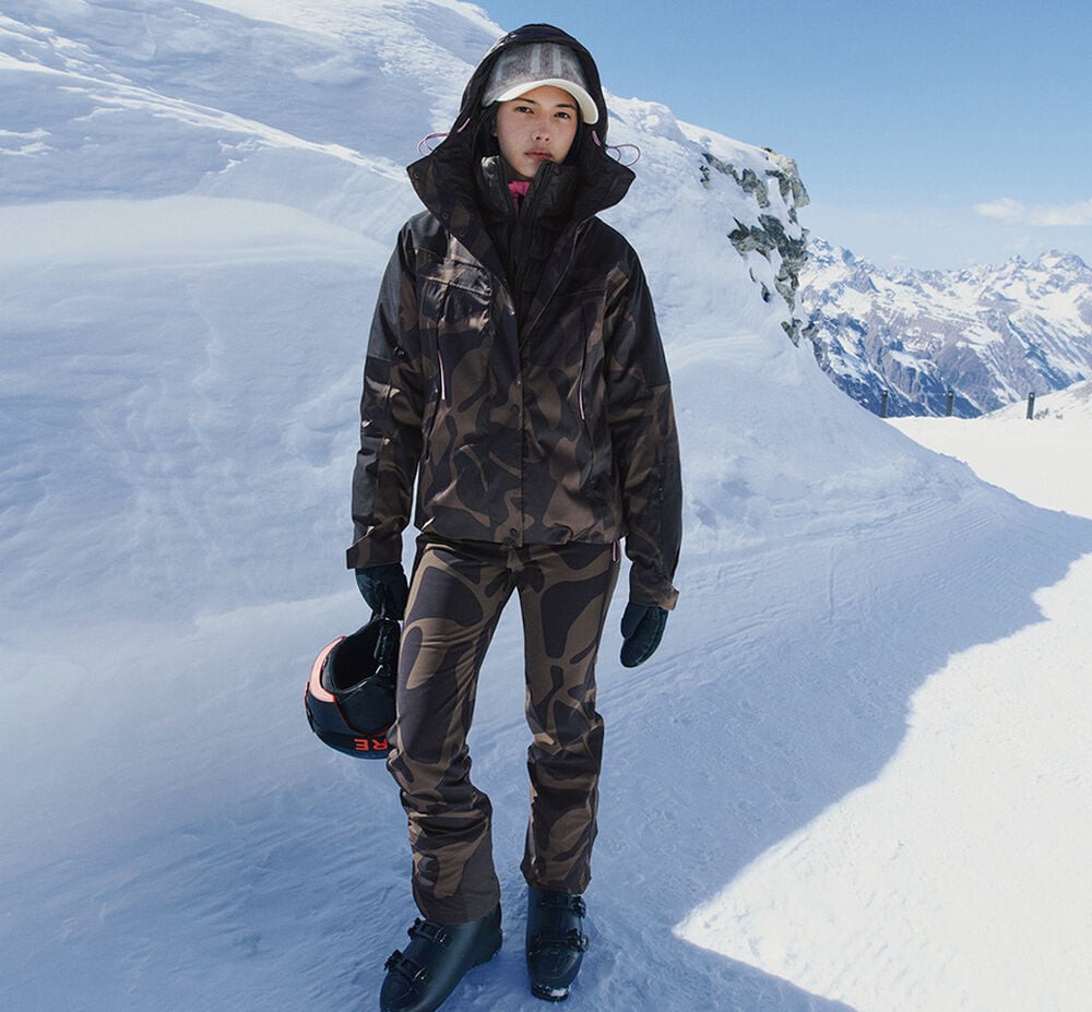 Das Damen Model trägt einen kompletten Ski Look mit Camouflage-Print, bestehend aus Jacke, Hose, schwarzem Helm und Skischuhen.