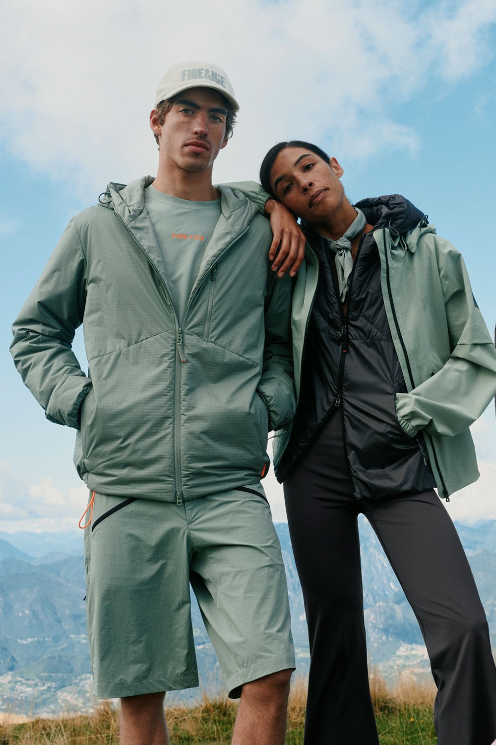 A male and female model wearing FIRE+ICE looks in eucalyptus and anthracite. They are standing on the grass with mountain scenery in the background.